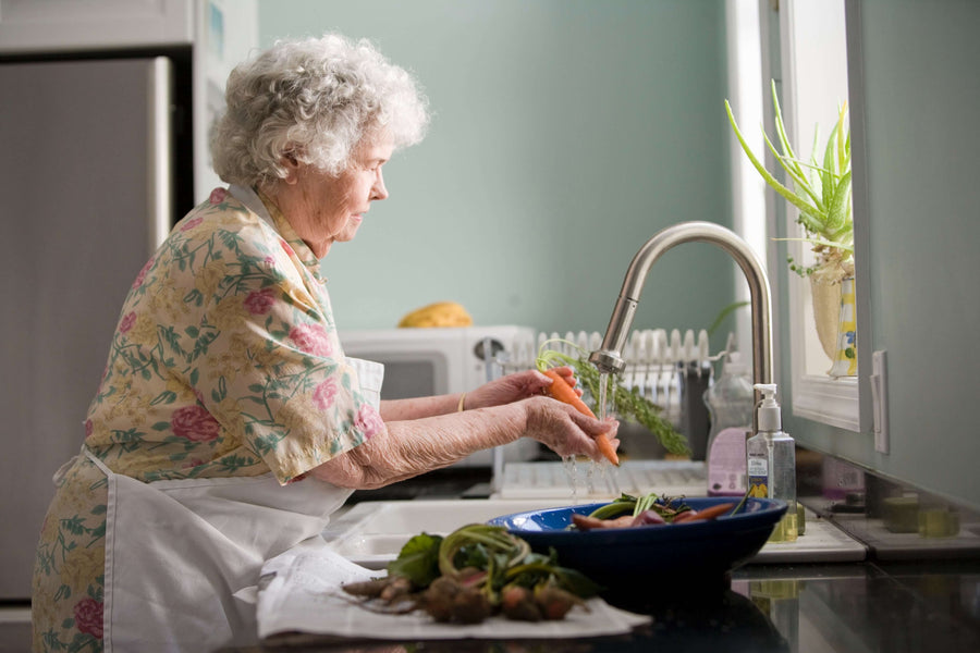 Older woman in apron washes vegetables at sink